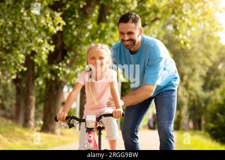 Père affectueux d'âge moyen et sa jolie fille passant du temps ensemble hors portes, papa enseignant à l'enfant comment faire du vélo Banque D'Images