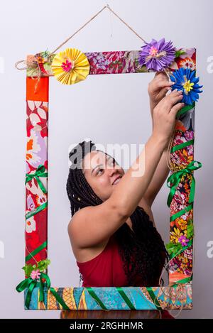 Belle femme derrière un cadre orné de fleurs et de tissus colorés en position verticale touchant les fleurs. Arrière-plan de couleur blanche. Banque D'Images