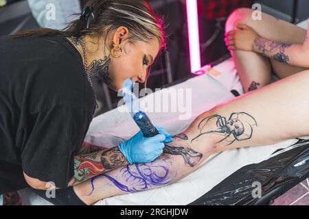 Femme tenant la machine de tatouage et dessinant un tatouage sur la jambe du client. Procédé de tatouage de veau. Artiste féminine concentrée pendant le travail. Photo de haute qualité Banque D'Images