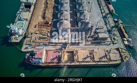 Chargement du grain dans les cales d'un navire de fret maritime dans le port maritime à partir de silos de stockage du grain. Soutage de cargaisons sèches avec du grain. Vue de dessus de l'antenne. Banque D'Images