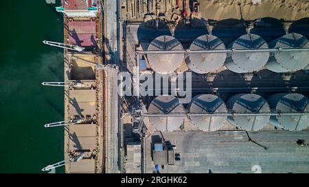 Chargement du grain dans les cales d'un navire de fret maritime dans le port maritime à partir de silos de stockage du grain. Soutage de cargaisons sèches avec du grain. Vue de dessus de l'antenne. Banque D'Images