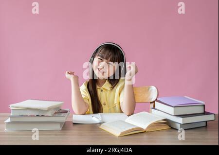 Une jeune fille asiatique adorable et heureuse aime écouter de la musique sur ses écouteurs à sa table d'étude sur un fond rose isolé. Banque D'Images