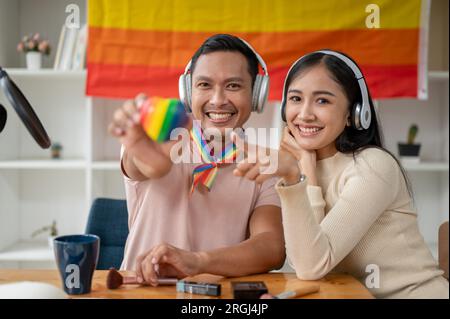 Un gay asiatique attrayant et heureux dans des écouteurs tenant le cœur LGBT dans sa chambre avec son amie. Diversité, homosexuel, gay, amitié, Banque D'Images