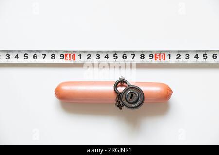 Saucisse sur une chaîne sous serrure et clé et à côté d'elle se trouve une bande centimétrique sur un fond blanc au soleil, le rejet de la nourriture malsaine, le régime alimentaire et OV Banque D'Images