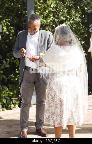 Heureux marié biracial senior mettant la bague sur le doigt de la mariée à la cérémonie de mariage en plein air ensoleillé Banque D'Images