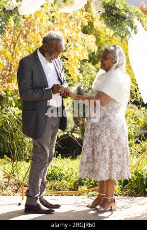 Heureux marié biracial senior mettant la bague sur le doigt de la mariée à la cérémonie de mariage en plein air ensoleillé Banque D'Images