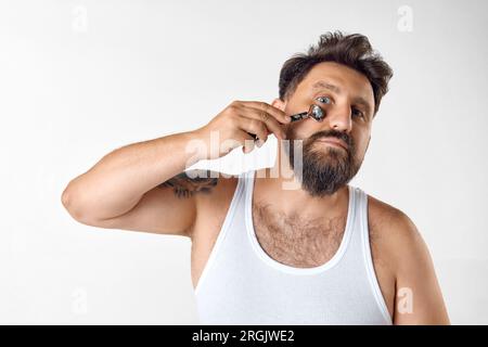 Rafraîchissement matinal. Homme barbu prenant soin après la peau, faisant massage du visage avec rouleau sur fond de studio blanc Banque D'Images
