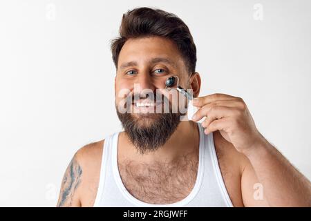 Souriant, homme barbu faisant massage du visage avec rouleau sur fond de studio blanc. Elimination des poches Banque D'Images