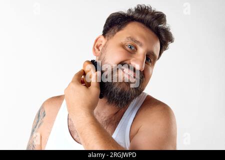 Mature, souriant, bel homme prenant soin après la barbe, brossant avec une brosse à cheveux spéciale sur fond de studio blanc Banque D'Images