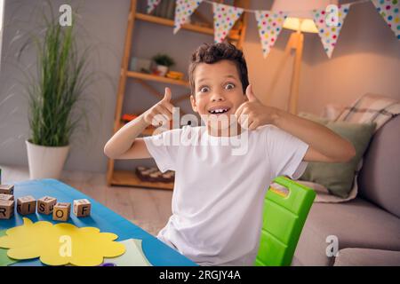 Photo de mignon garçon émerveillé assis bureau préscolaire Profitez de l'activité extrascolaire montrer le symbole pouce vers le haut dans la salle de jeux confort Banque D'Images