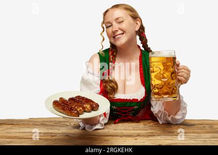 Belle rousse fille dans la tenue folklorique bavaroise clin tenant des tasses remplies de bière et assiette avec des saucisses. Portrait de l'Oktoberfest Banque D'Images