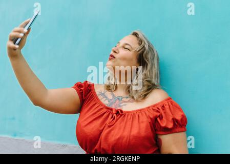 Vue latérale de jeune femme de taille plus dans des vêtements décontractés prenant selfie dans la bouche ronde pose avec smartphone tout en se tenant sur fond de mur bleu à l'extérieur Banque D'Images