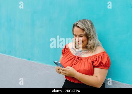 Angle élevé de jeune femme souriant et regardant smartphone tout en se tenant sur la rue de la ville sur fond bleu Banque D'Images