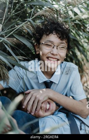 Heureux garçon asiatique en uniforme scolaire avec les cheveux bouclés et les lunettes souriant et jouant avec le jeune frère tout en regardant la caméra et se reposant par le vert Banque D'Images