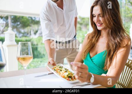 Assiette de serveur méconnaissable avec salade savoureuse avec crevettes et légumes pour la cliente assise à table dans le patio du restaurant pendant lu Banque D'Images