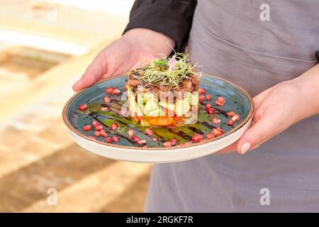 Récolte méconnaissable chef féminin en tablier debout avec une assiette de tartare délicieux fait de poisson avec avocat et tomates Banque D'Images