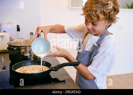 Vue de côté de la culture souriant enfant ajoutant de la crème de lait aux tranches de bacon dans la poêle tout en préparant la sauce carbonara dans la cuisine Banque D'Images