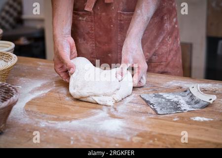 Les mains du boulanger mâle travaillent de la pâte à pain avec de la farine sur une table en bois. Banque D'Images