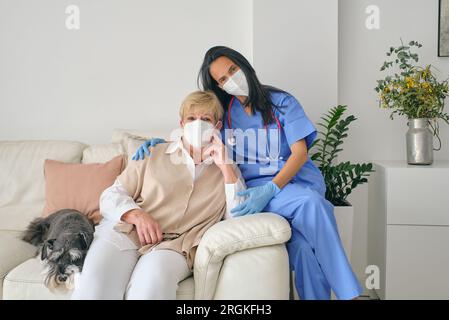 Femme médique en uniforme et masque respiratoire enveloppant le patient senior sur le lit avec Schnauzer miniature tout en regardant la caméra Banque D'Images