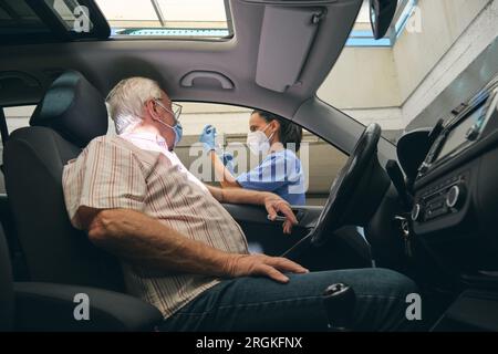 Patient mâle âgé méconnaissable en automobile contre une femme médecin en masque respiratoire avec injecteur et vaccin COVID 19 Banque D'Images