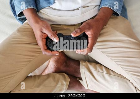 D'en haut de la récolte anonyme joueur masculin afro-américain portant une tenue décontractée jouant à un jeu vidéo avec manette de jeu tout en étant assis sur un canapé doux avec des jambes cros Banque D'Images