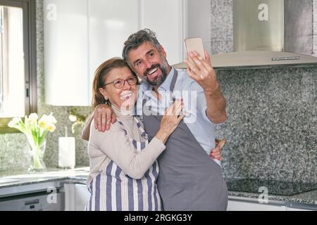 Mère aînée positive dans les lunettes et fils joyeux non rasé dans les tabliers serrant dans la cuisine et prenant l'autoportrait tout en regardant l'écran de smartp Banque D'Images