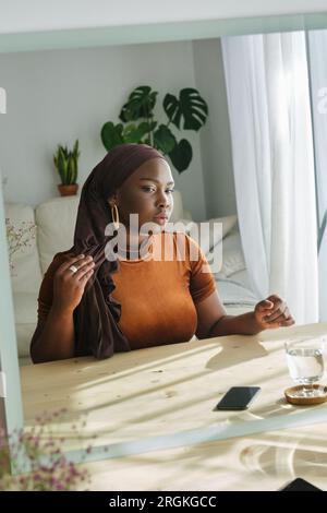 Reflet de femme noire musulmane pensive en tenue tendance et foulard assis à la table en bois et s'admirant dans le miroir dans le salon sur le soleil Banque D'Images