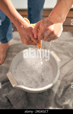 Du dessus de la personne sans visage en jeans debout sur la serviette et essorant le chiffon sur le bassin blanc avec de l'eau mousseuse tout en faisant la routine domestique à la maison Banque D'Images