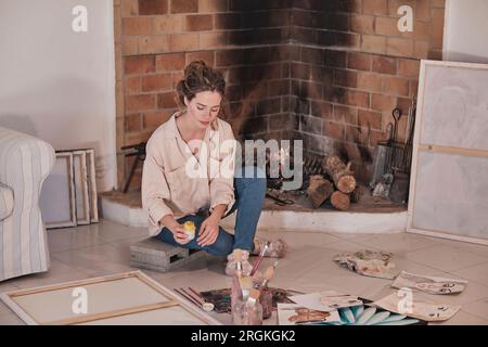 Artiste féminine ciblée dans des vêtements décontractés assis sur le sol près de la cheminée et dessiner en utilisant des peintures professionnelles dans l'espace de travail créatif Banque D'Images