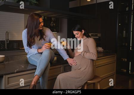 Femme positive avec mug touchant le ventre de l'amie enceinte et souriant tout en étant assis sur la table en marbre dans la cuisine confortable à la maison le week-end Banque D'Images
