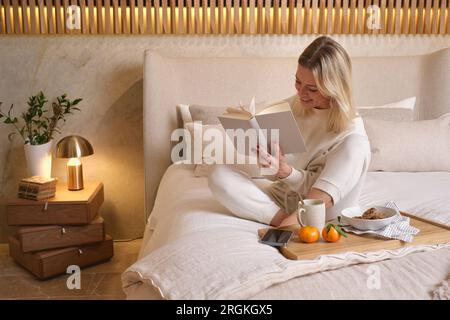 Corps entier de femme souriante en vêtements de nuit blancs assis sur un lit doux près d'un plateau en bois avec petit déjeuner et lecture d'un livre intéressant dans la chambre à la maison Banque D'Images