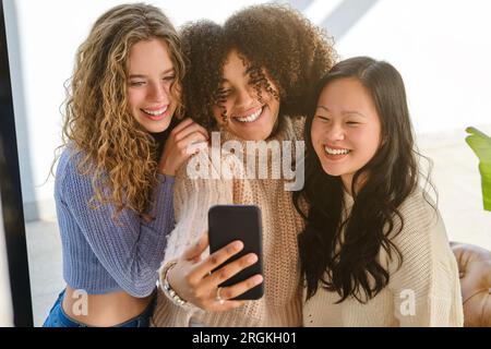 Heureux jeunes femmes diverses se tenant proches les uns des autres souriant et prenant selfie sur smartphone Banque D'Images