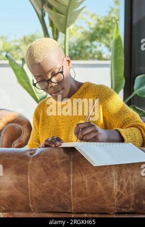 Concentré jeune femme afro-américaine écrivant des notes dans le journal assis sur le canapé à la maison Banque D'Images