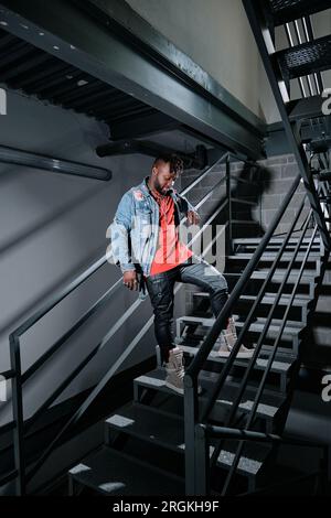 Jeune homme noir dans les vêtements de style de rue pensivement debout sur l'escalier dans le bâtiment et regardant vers le bas Banque D'Images