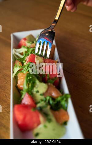 Photo verticale d'une main d'une personne mangeant un saumon mariné avec une salade d'escarole et une sauce moutarde à la fourchette et servi dans une coupe rectangulaire blanche Banque D'Images