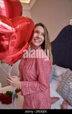 Femme joyeuse sur le lit avec un bouquet de ballons en forme de coeur rouge reposant dans la chambre d'hôtel le matin de vacances Banque D'Images