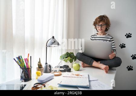 Corps entier de l'enfant pieds nus avec les cheveux bouclés surfant sur les médias sociaux sur netbook et regardant l'écran tout en étant assis près de la fenêtre à la maison Banque D'Images