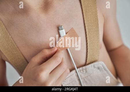 D'en haut, récolte anonyme enfant en beige collant le chargeur de téléphone sur la poitrine avec du plâtre dans la pièce lumineuse Banque D'Images