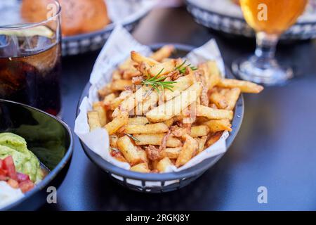 Angle élevé de frites savoureuses surmontées de romarin et servies dans un panier noir sur table avec divers plats et de la bière dans le café de rue Banque D'Images