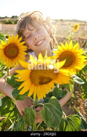 Enfant souriant avec les cheveux bouclés debout au milieu des tournesols en fleurs sur le champ de campagne et regardant la caméra sur la journée ensoleillée Banque D'Images