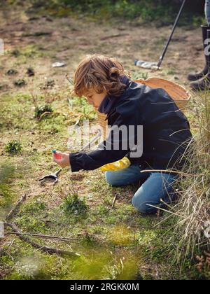 Vue latérale du garçon préadolescent concentré assis sur les genoux et ramasser les déchets plastiques et les ordures sur un sol herbeux en journée ensoleillée Banque D'Images