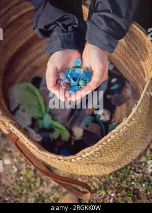 Vue de dessus des mains de la culture anonyme enfant tenant des morceaux de plastique pour le recyclage sur le grand sac en osier pour collecter les déchets en plastique à l'extérieur sur le sol Banque D'Images