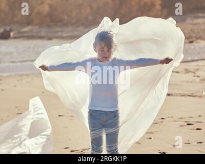 Garçon préadolescent paisible debout avec les mains tendues de côté enveloppé dans un film plastique ramassé du rivage sablonneux contaminé de rive dans la journée ensoleillée Banque D'Images