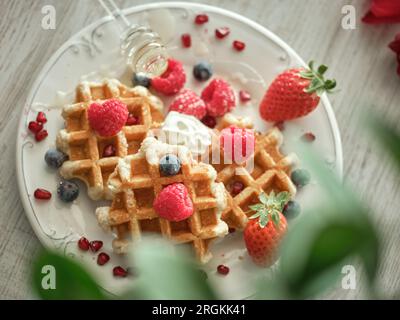 D'en haut de gaufres sucrées servies sur une assiette avec des baies mûres et une trempette de miel dans une cuisine légère avec une plante verte floue Banque D'Images