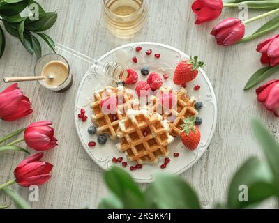 Gaufres décorées de diverses baies sur l'assiette contre des tulipes rouges avec pot de miel et verre de café sur la table Banque D'Images