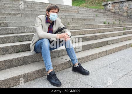 Un employé de bureau portant un masque médical, licencié de son travail, est assis sur les marches de la rue. Banque D'Images