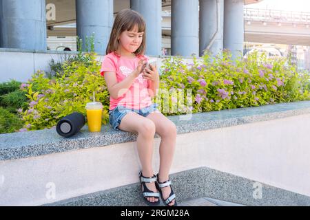 Belle petite fille est assise dans la rue et regarde le téléphone portable Banque D'Images