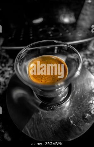 Vue macro d'une tasse en verre avec du café noir fraîchement préparé avec de la mousse dorée avec de petites bulles dans une soucoupe à café en métal sur le dessus d'un barista automatique Banque D'Images