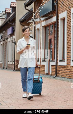 heureuse femme âgée tenant une tasse de papier et marchant avec des bagages, café à emporter, mode de vie urbain Banque D'Images