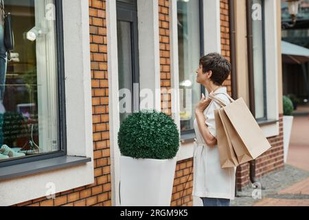 femme âgée avec les cheveux courts tenant des sacs à provisions et regardant la fenêtre du centre commercial extérieur, outlet Banque D'Images